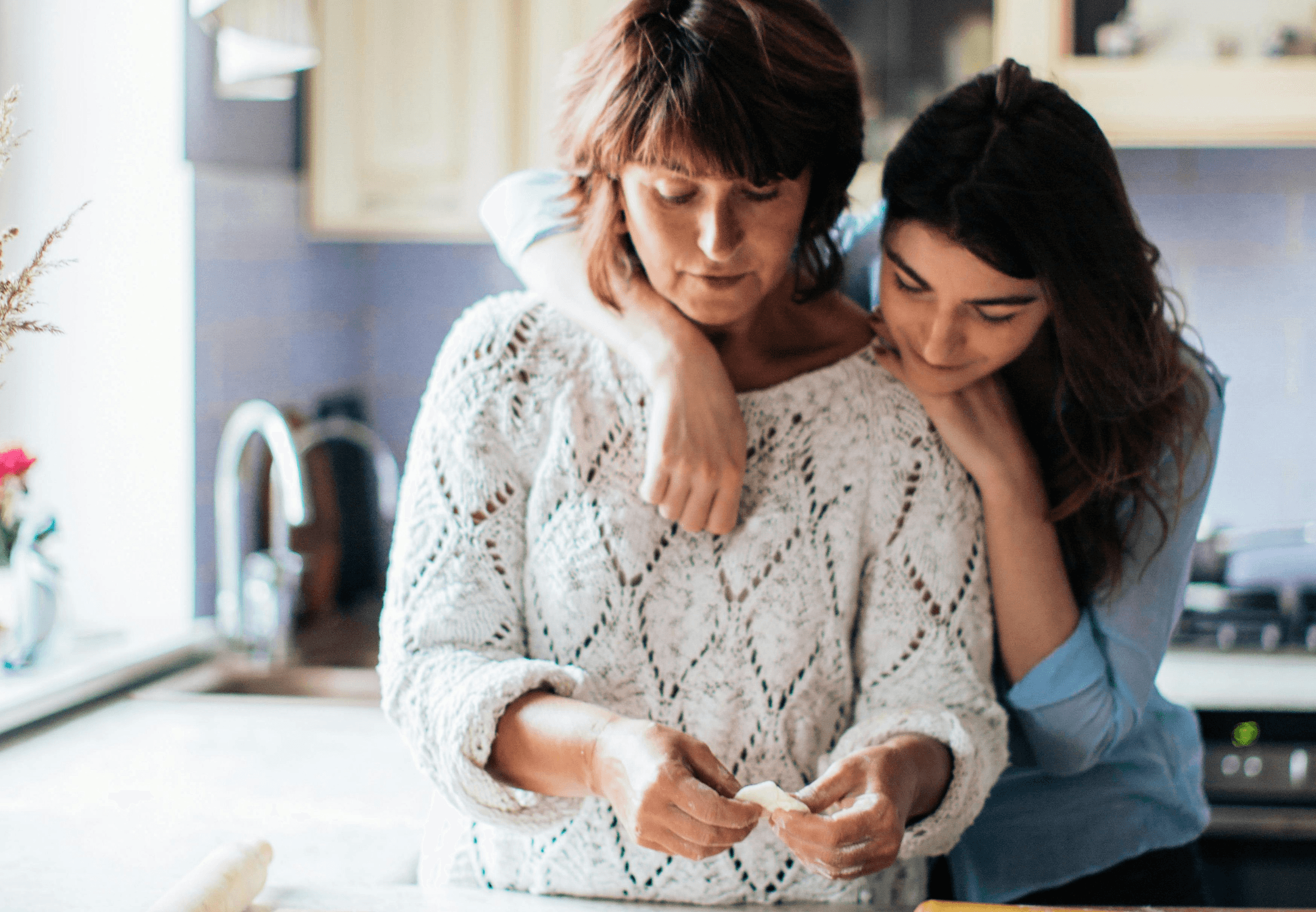 A mother and daughter stand together in a loving embrace while the mother prepares food on the kitchen bench. 